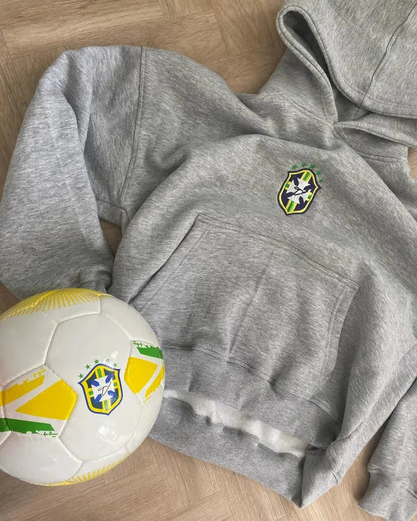 Grey BRASIL pullover hoodie with a crest detail and front pouch pocket, styled beside a football